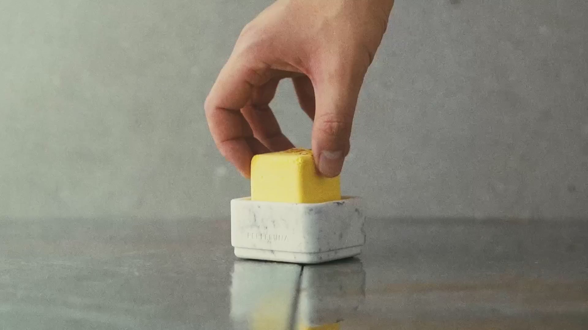 Ruka vkládá žlutou sprchovou aromatickou kostku do bílé kamenné misky, pak na ni teče voda